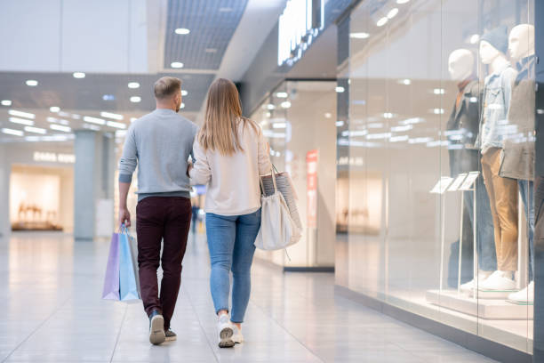 vista posteriore della giovane coppia casual che si muove lungo la vetrina del centro commerciale - guardare le vetrine foto e immagini stock