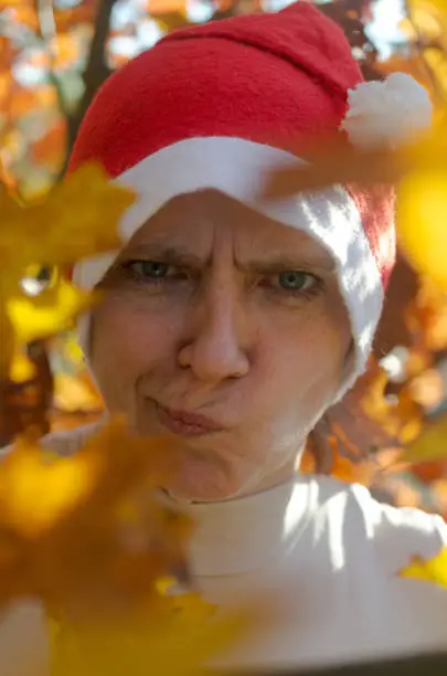 A woman in a hat of Santaclaus with a sly evil face among the autumn foliage as a harbinger of the coming winter