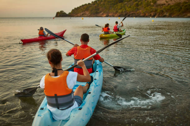 Lens flare view of Spanish couples in 30s and 40s paddling kayaks in Mediterranean Sea as sun rises over Costa Brava headland in background.