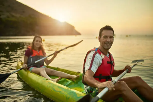 Close-up of active Spanish friends in late 20s and mid 40s paddling their tandem kayak off the Costa Brava with sun coming up over headland in background.