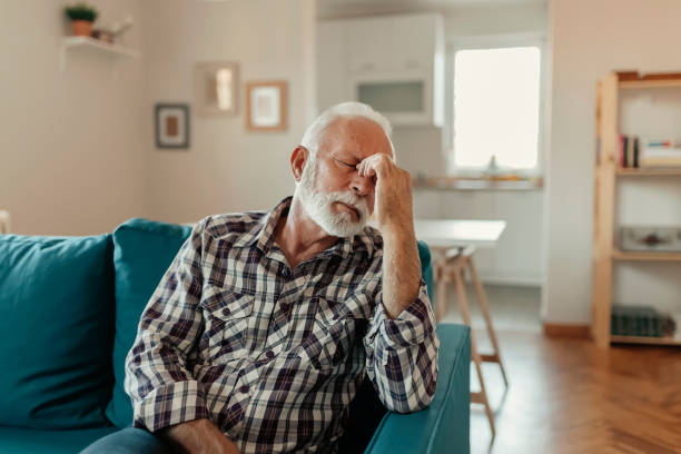 This Headache! A Distraught Senior Man Suffering From a Migraine While Sitting on the sofa in the Living Room gurning stock pictures, royalty-free photos & images