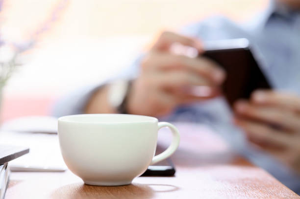 istock White coffee cup on wooden table in cafe with lighting and blurred image man using smartphone background. 1185130574