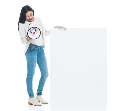 Full length / one person / front view of 20-29 years old adult beautiful latin american and hispanic ethnicity female / young women standing in front of white background wearing blazer - jacket / jacket / jeans who is talking / screaming / shouting / smiling / happy / cheerful who is showing and holding sign / megaphone and using placard with copy space