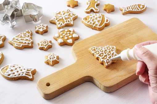 Women hand using a pastry bag to decorate gingerbread cookies with patterns from royal icing for Christmas, kitchen board on a white painted table, selected focus, narrow depth of field