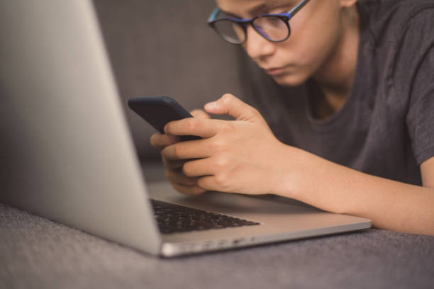 istock Young student doing homework lying on a sofa using laptop and smartphone. Caucasian boy communicate with friends. Teen enjoying  new electronic devices. Technology education concept Soft focus on hand 1185191144