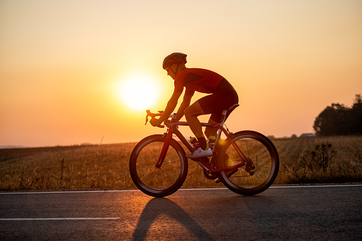 Young professional athlete riding bicycle for exercise on the road through the countryside. He is well equipped, with protective helmet, sunglasses, black and red jersey. Photo taken when cyclist is in motion. Sunset in background makes him silhouette.