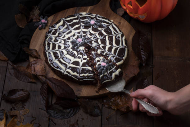 Spiderweb cake made of sticky chocolate cake kladdkaka with a Halloween pumpkin in the background. A hand is taking a slice of cake. The cake is on a wooden table, and is surrounded by autumn leaves.