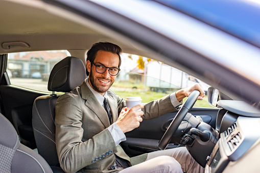 Handsome man driving a car and holding paper cup of coffee while looking away. Photo Of Business Man Driving Car. Succesful Young Smiling Businessman Drinking Coffee From Paper Cup While Driving Car. Handsome Happy Male Going To Work In Comfortable Car.