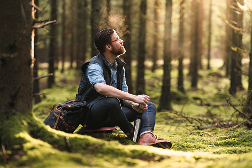 A man hiking through pine tree forests in Sweden. He is sitting down to drink some hot coffee.