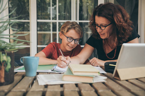 istock Young student doing homework at home with school books, newspaper and digital pad helped by his mother. Mum writing on the copybook teaching his son. Education, family lifestyle, homeschooling concept 1185202596