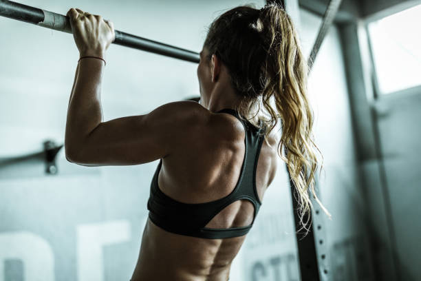 Rear view of athletic woman exercising chin-ups in a gym. Back view of determined female athlete exercising chin-ups during cross training in a health club. chin ups photos stock pictures, royalty-free photos & images