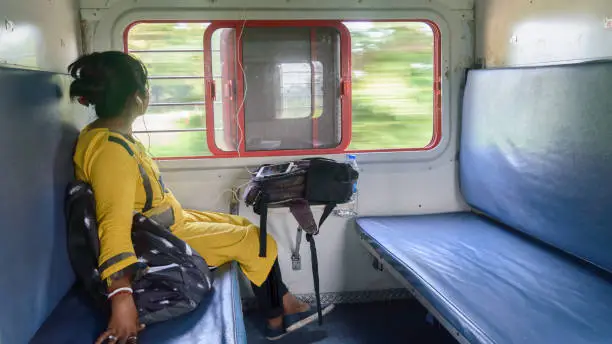 Woman listening music on mobile phone and looking outside from window while travelling solo in train. Traveler enjoying Modern technology on the move in everyday life and travel. Side view Close up portrait - Young Adult Lady - Indian ethnicity.