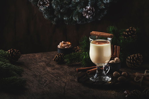 New Year or Christmas Eggnog cocktail - hot winter or autumn drink with milk, eggs and dark rum, sprinkled with cinnamon and nutmeg in a glass on wooden background, festive decoration