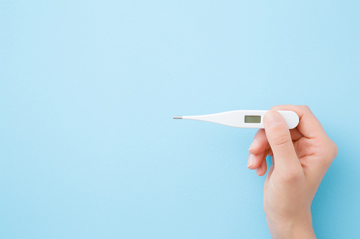 Young woman hands holding white digital thermometer on pastel blue background. Fever and healthcare concept. Closeup. Point of view shot. Empty place for text. Top down view.