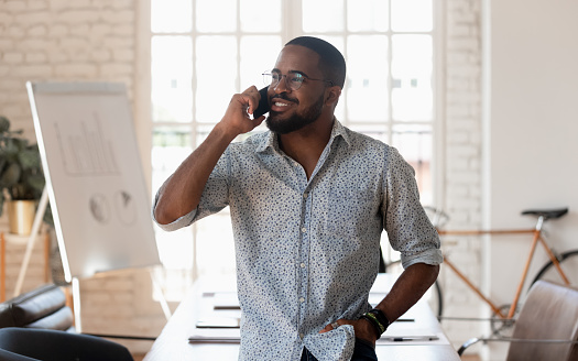 Successful millennial mixed race businessman standing at creative modern office, holding mobile phone call with potential clients, communicating negotiating with partners, listening to good news.