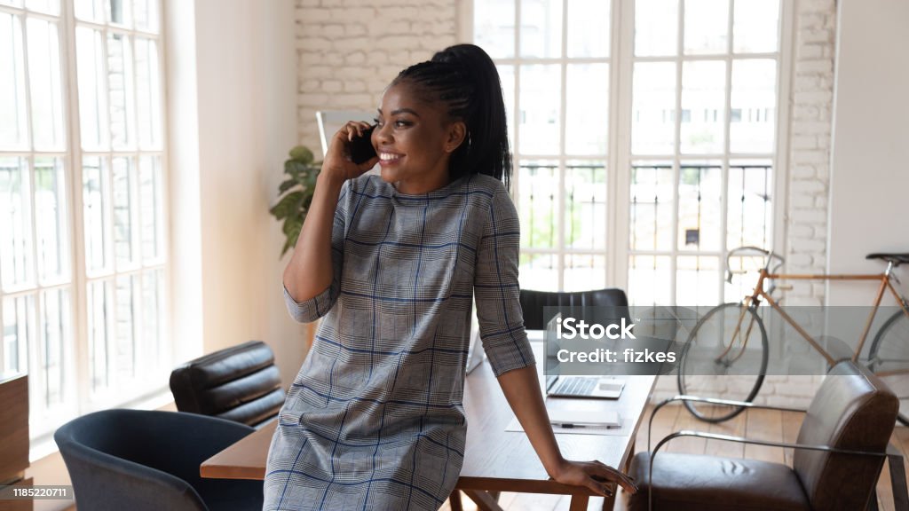 Happy millennial african american female manager talking on mobile phone. Happy millennial african american female manager standing at modern creative office, talking on mobile phone with partners, negotiating with clients, consulting customers or solving business issues. Women Stock Photo