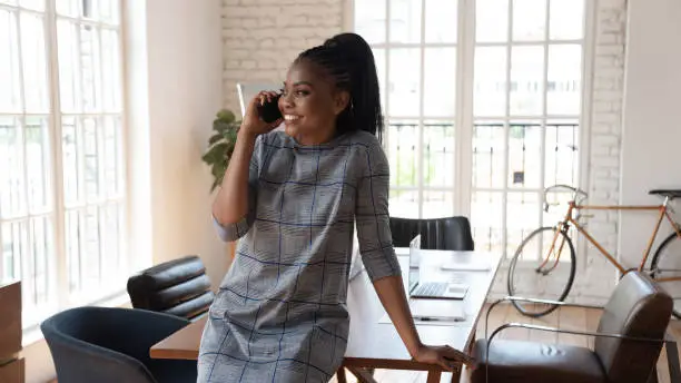 Happy millennial african american female manager standing at modern creative office, talking on mobile phone with partners, negotiating with clients, consulting customers or solving business issues.