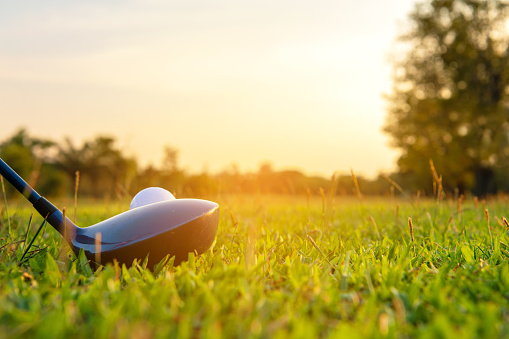 Cerrar la pelota de golf y el conductor, jugador haciendo golf swing tee fuera en la hora verde de la noche de puesta de sol, presumiblemente hace ejercicio.  Concepto de estilo de vida y salud. photo