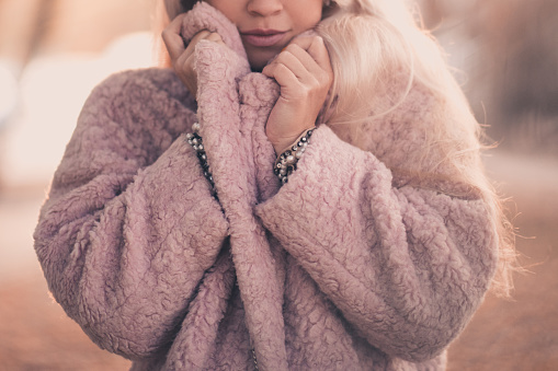 Beautiful woman wearing cozy pink fur coat outdoors over sunny background in city. Winter season.