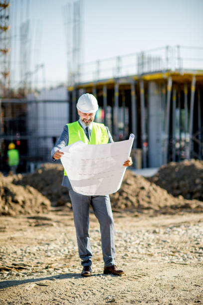istock Cheerful positive handsome senior engineer  wearing hardhat satisfied with building plan holding blueprint while working at construction site 1185239325
