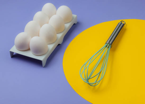 istock Tray with white chicken eggs and whisk on blue background with yellow circle in the middle. Minimalistic food concept. 1185251204