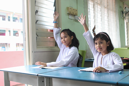 Two Malay schoolgirls raising their hands in a classroom to answer a question.