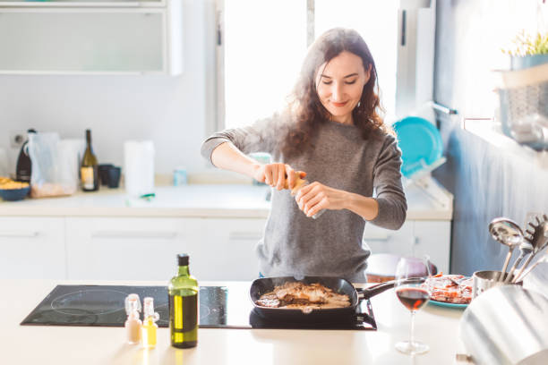 台所で調理する若い笑顔の女性は、焼き肉にコショウを加えます。健康的な食品のコンセプト。 - 女性料理人 ストックフォトと画像