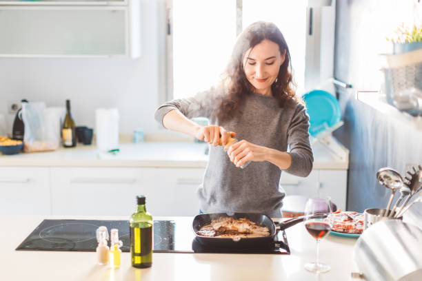 jonge lachende vrouw koken in de keuken, het toevoegen van peper aan het gegrilde vlees. gezond voedsel concept. - barbecue maaltijd fotos stockfoto's en -beelden