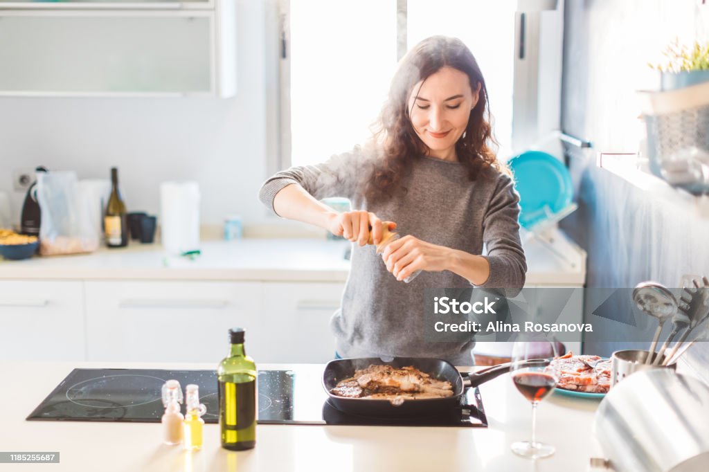 Young smiling woman cooking in the kitchen, adding pepper to the grilled meat. Healthy food concept. Picture of woman grilling meat Cooking Stock Photo