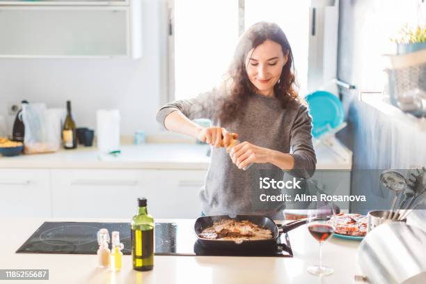 Young Smiling Woman Cooking In The Kitchen Adding Pepper To The Grilled Meat Healthy Food Concept Stock Photo - Download Image Now