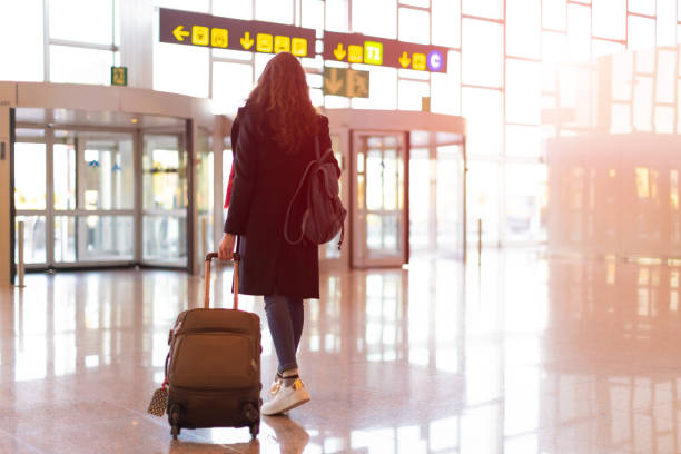 rückansicht der brünetten frau, die mit trolley vom flughafen aussteigt (handgepäck) - airport lounge airport one person bag stock-fotos und bilder