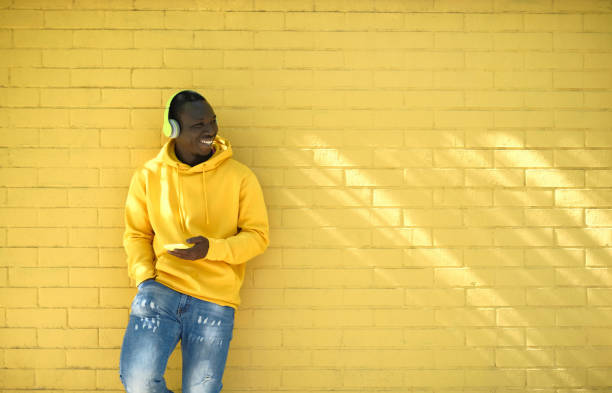 African Young Man Listening Music Leaning On Yellow Wall Young African Man With Wireless Headphones Streaming Music On Telephone, Leaning Against Yellow Wall And Smiling While Looking Away. jersey fabric photos stock pictures, royalty-free photos & images