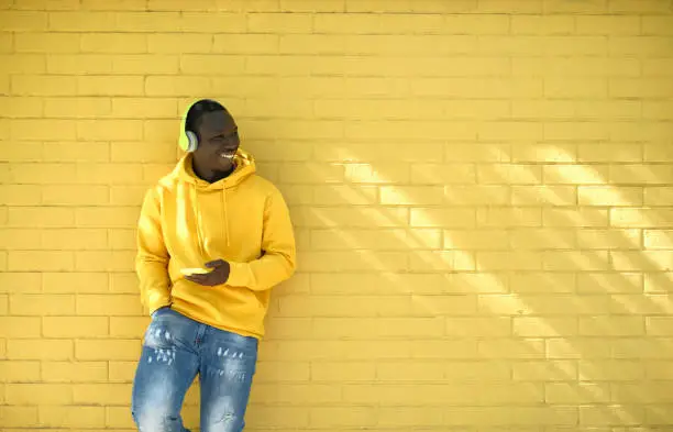 Young African Man With Wireless Headphones Streaming Music On Telephone, Leaning Against Yellow Wall And Smiling While Looking Away.