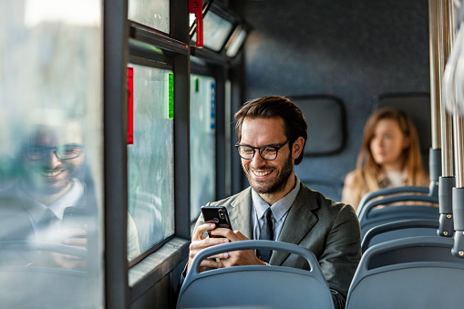 Smiling Confident young businessman in full suit commuting to work by bus and having a phone call with his smartphone