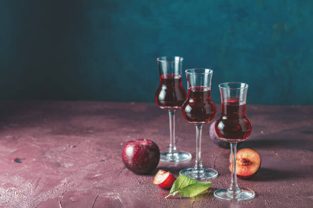 plums strong alcoholic drink in grappas wineglass with dew. hard liquor, slivovica, plum brandy or plum vodka with ripe plums on dark blue and  claret bordeaux concrete surface - plum fruit organic food and drink imagens e fotografias de stock