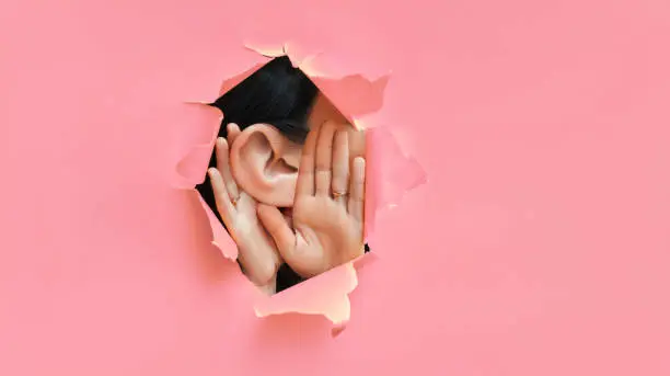 Photo of Female ear and hands close-up. Torn paper, pink background. The concept of eavesdropping, espionage, gossip and the yellow press. Caricature with an enlarged ear.