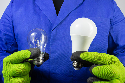 Photo of electrician in blue robe and gloves holding old bilb and new led light bulb close-up view.