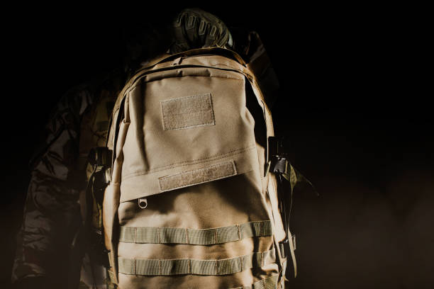 Photo of a fully equipped soldier in uniform holding backpack. Photo of a fully equipped soldier in uniform, armor and mask holding backpack on dark background with fog and dust close-up view. khaki green photos stock pictures, royalty-free photos & images