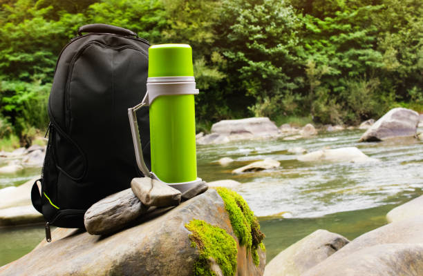 istock Travel backpack with thermos bottle standing on forest river rocks. 1185310965