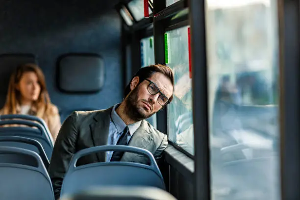 Tired young businessman sitting on the bus and day dreaming while leaning on the window during the day.