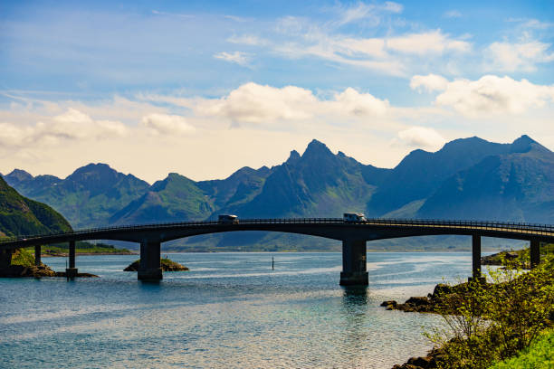 Norwegian scenic landscape on Lofoten archipelago. Camper vehicle on road and bridge connecting islands over sea. Mountain peaks in distance.