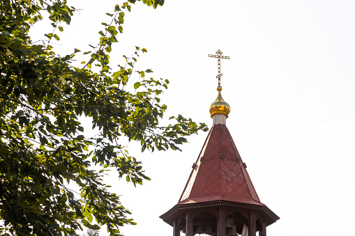 Maroon top of orthodox church bell chamber with cross against sky and tree leaves