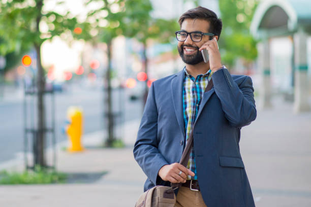 Walking and Talking An attractive Indian businessman is talking on his smartphone while walking to work downtown. He smiles and wears a suit jacket. indian man walking in park stock pictures, royalty-free photos & images