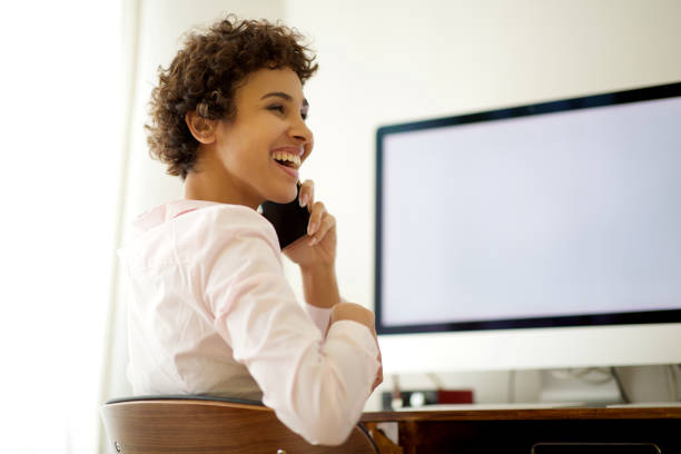 Portrait of smiling young woman talking with mobile phone by computer screen