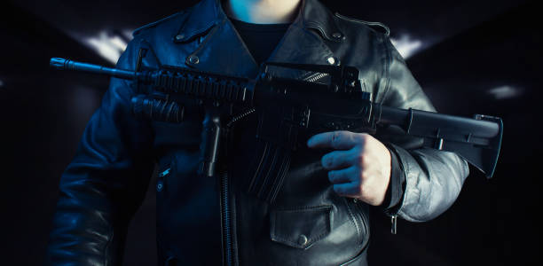Biker in black leather jacket with skull rings holding rifle.