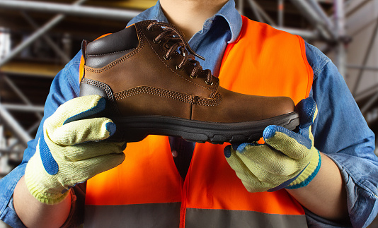 Worker in orange jacket outfit in protective gloves holding leather boot on construction area background.