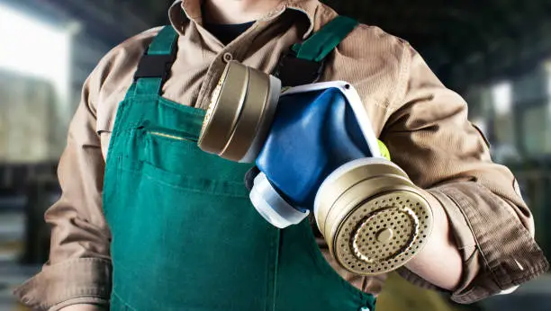 Photo of a worker in green overall outfit with protective gloves holding blue filtered respirator on factory background torso view.