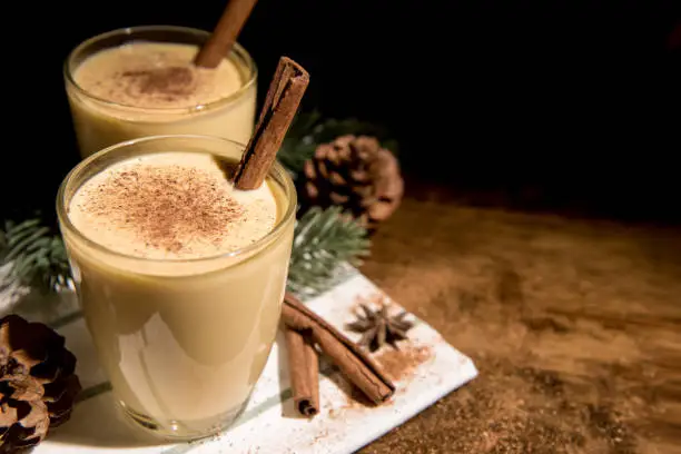 Homemade traditional Christmas eggnog drinks the glasses with ground nutmeg, cinnamon and decorating items on wood table, preparing for celebrating festive holiday season