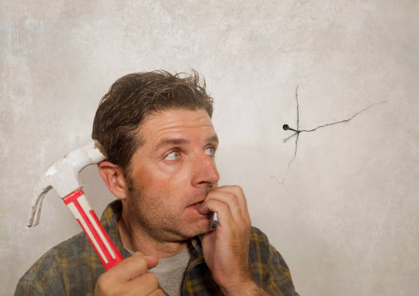 funny portrait of man holding hammer after driving a nail for hanging a frame but making a mess cracking the wall as a disaster diy guy and messy domestic repair task - home improvement work tool hammer portrait imagens e fotografias de stock