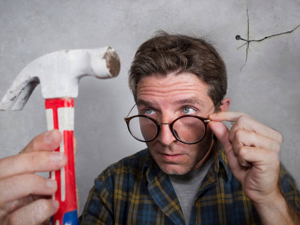 portrait of nerd man looking to hammer after driving a nail for hanging a frame making funny face for the  mess cracking the wall as a disaster diy guy and messy domestic repair task at home - home improvement work tool hammer portrait imagens e fotografias de stock